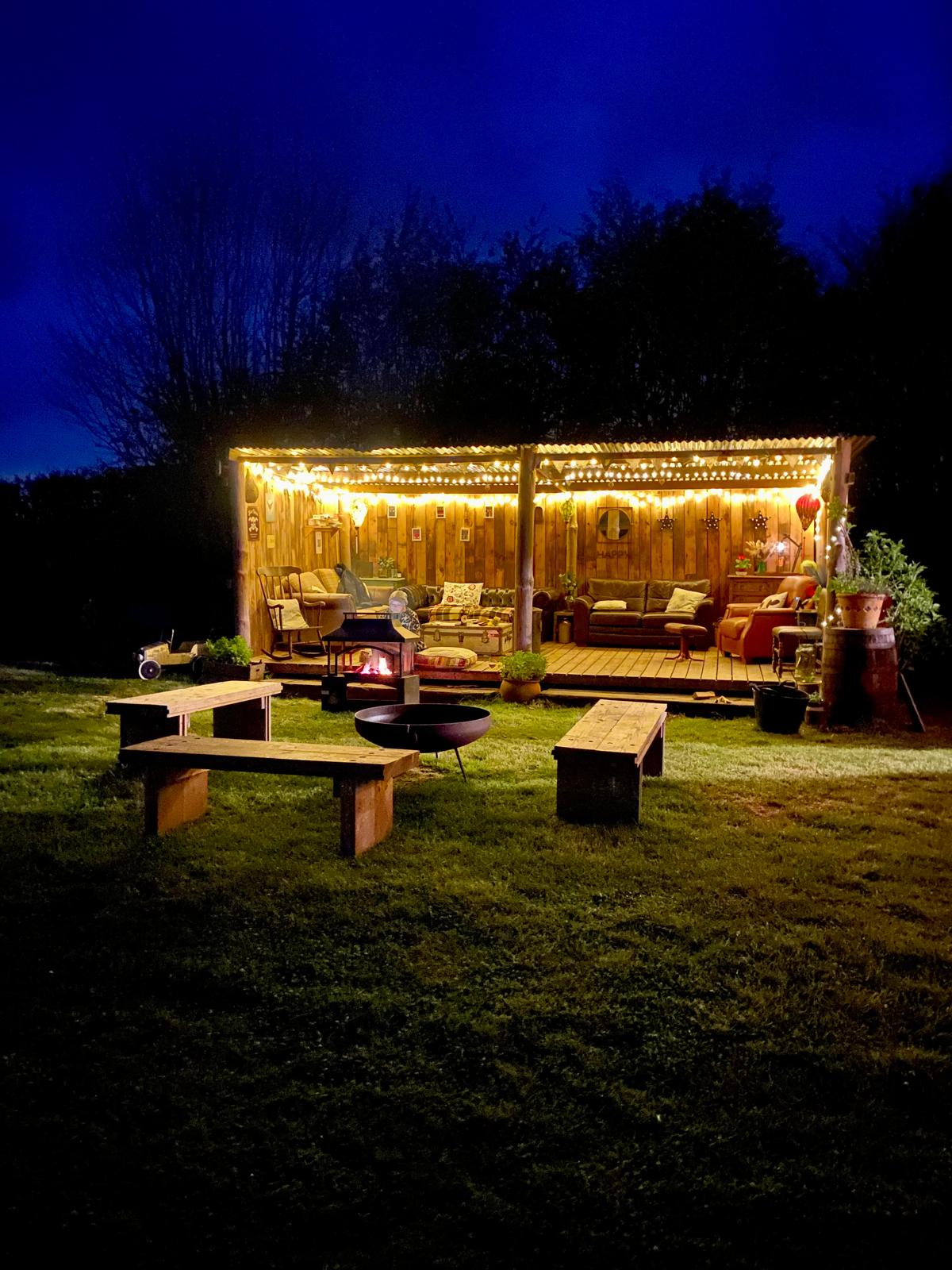 The wooden barn lit with fairy lights at dusk with fire pit and benches